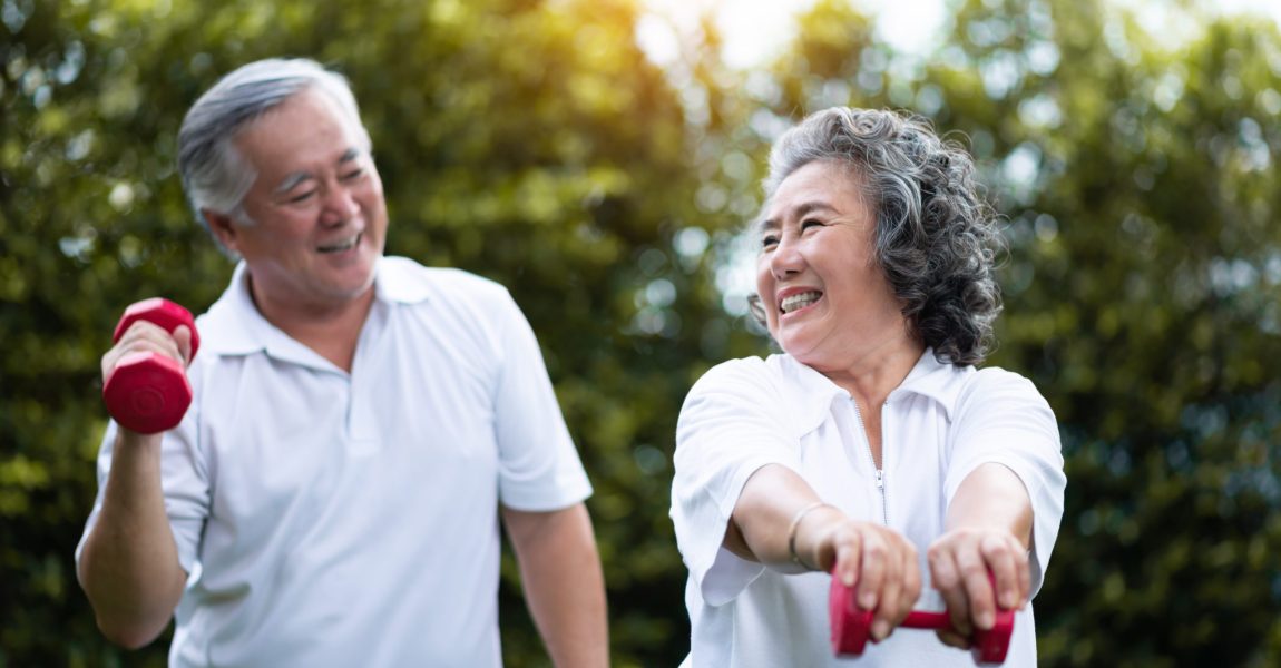 Asian Senior Couple exercising with red dumbbells at the outdoor park togetherness. Smiling Chinese or Thai or Japanese people.